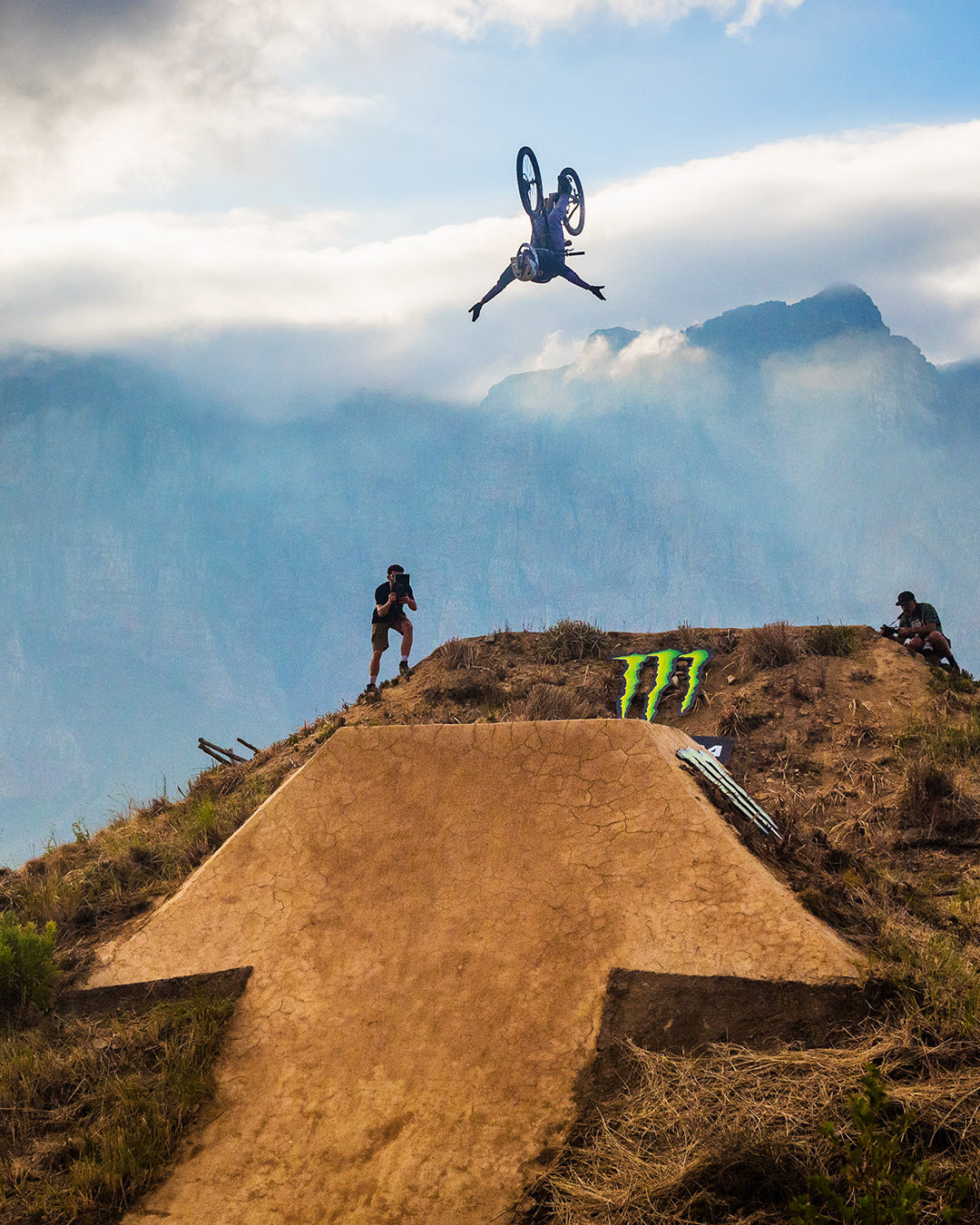 Person on a bicycle with Kenda tires performing a jump over a dirt ramp with mountains in the background