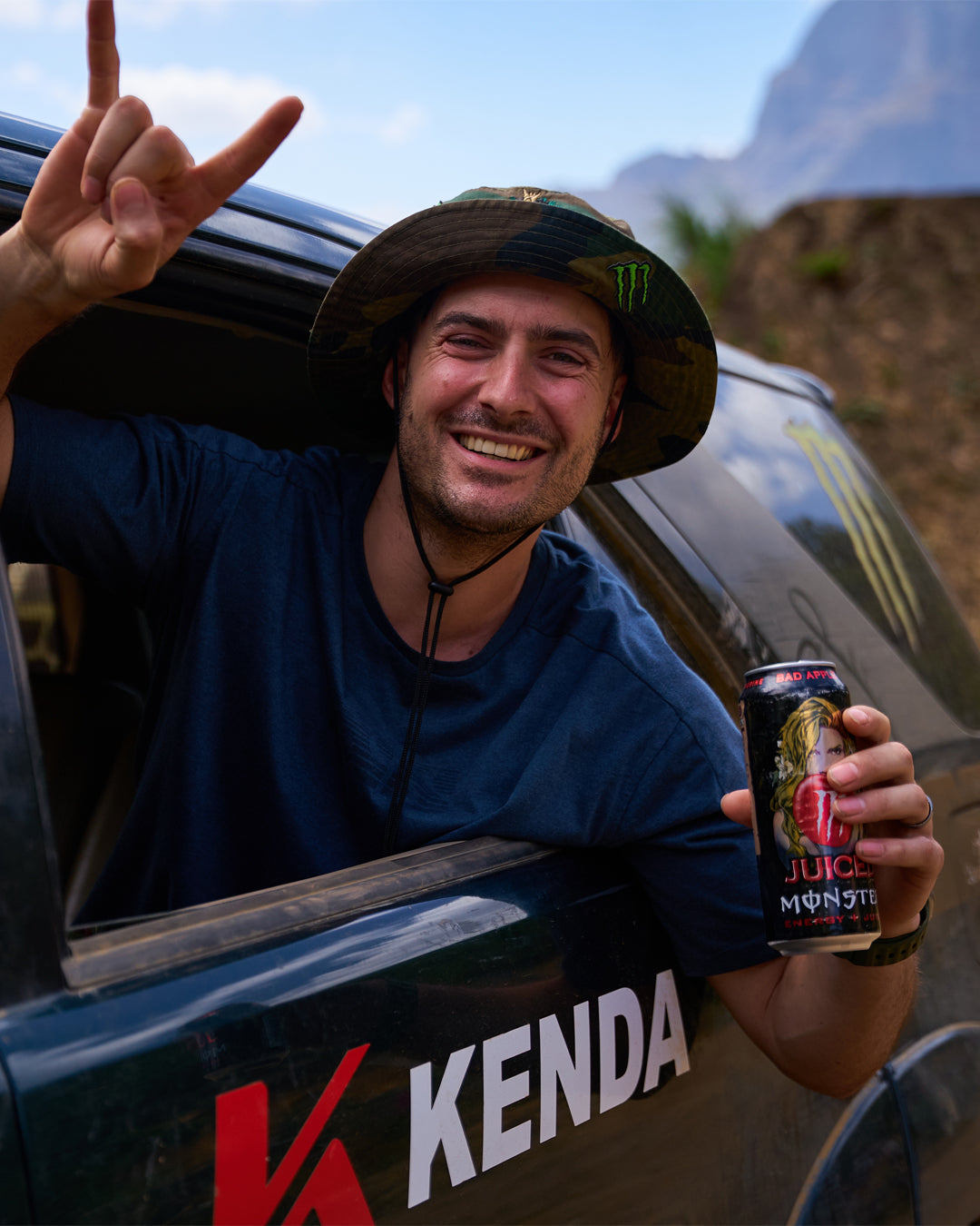 Man in a blue shirt and hat holding a can of Monster Energy drink, sitting in a vehicle with Kenda branding.